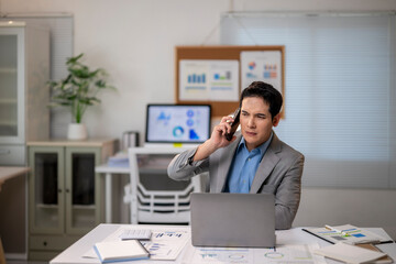 A man in a suit is talking on his cell phone while sitting at a desk with a lapt
