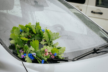 A handmade floral wreath of oak leaves and wildflowers lies on the windshield of a white car,...