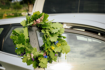 A bouquet of fresh oak leaves and wildflowers decorates the top corner of a car window, symbolizing...