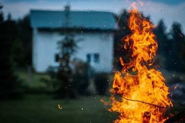 Bright orange flames rise from a bonfire in the foreground, with a blurred house and dark trees in...