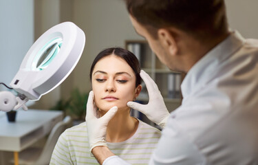Dermatologist doctor looking closely at woman face skin doing examination with dermatoscope...