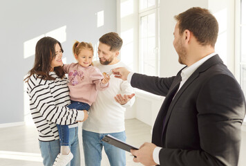 Portrait of man realtor giving house keys to a happy smiling young family of mother, father and...