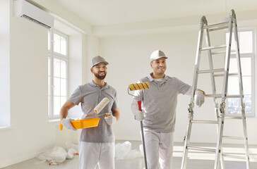 Two smiling male painters in uniform holding paint rollers and tray, standing in bright room near...