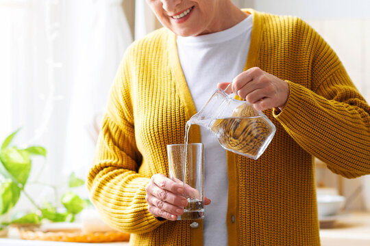 Hydration in senior ages concept. Unrecognizable grandmother standing in cozy white kitchen, pouring water into glass. Cropped of senior woman having healthy lifestyle, drinking water at home - Powered by Adobe