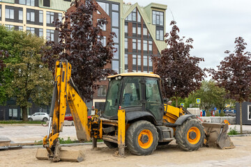 Yellow backhoe loader parked at urban construction site with modern buildings and trees in...