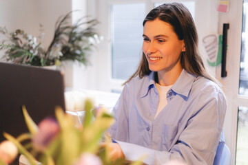 Smiling young woman sit at desk work on laptop or watching webinar, happy girl feel excited browsing internet on computer, using wireless home connection. Technology concept, girl wear blue shirt.