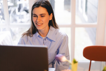 Smiling young woman sit at desk work on laptop or watching webinar, happy girl feel excited browsing internet on computer, using wireless home connection. Technology concept, girl wear blue shirt.