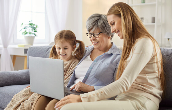 Grandmother, mother and child girl sitting on sofa using laptop together having online video call or watching movie. Smiling happy family resting on couch at home. People and technology concept.