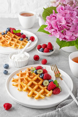 Delicious Belgian Waffle with whipped cream, fresh berries and mint on a white plate on a gray concrete background. Summer breakfast.