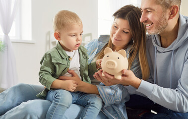 Happy young mother, father and adorable little son holding toy pink piggy bank, caring mum and dad...