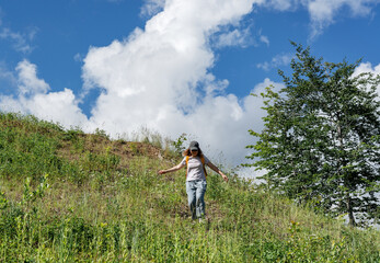Happy young woman hiker walking down a grassy hill under a bright blue sky with fluffy clouds, surrounded by vibrant greenery, enjoying a sunny day in nature, embodying freedom and joy