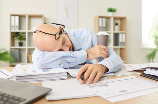 Overworked tired young business man putting head on table with a pile of financial documents and sleeping on workplace in office holding cup of coffee. Overload, overwork and stress concept.