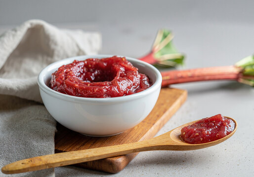 delicious homemade rhubarb jam in a white bowl garnished with fresh rhubarb stalks and accompanied by a wooden spoon on a rustic wooden cutting board