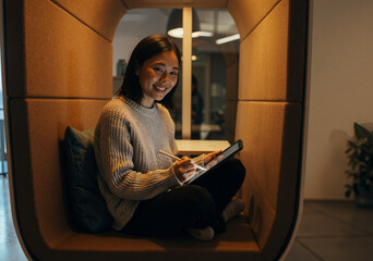 young woman sitting comfortably in a cozy workspace, using a tablet to design and create art, surrounded by modern and inviting decor in soft lighting
