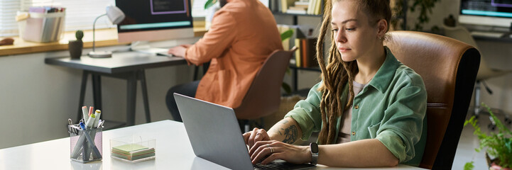 Young adult Caucasian woman developing neural network on laptop, sitting at desk in modern office,...
