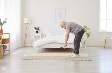 Senior elderly man wearing sleepwear standing in white bedroom near his bed and doing stretching...