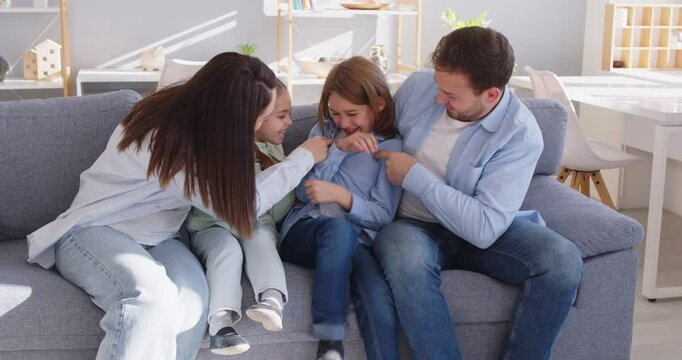 Happy family parents and children sitting on sofa together, having fun and fooling around. Joyful father, mother and daughter tickling laughing boy, playing carefree in living cozy room at home.
