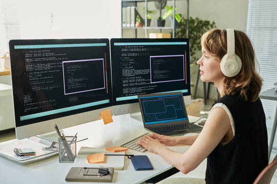 Caucasian young adult woman wearing headphones working on neural network development using dual monitors displaying code in modern office setting, typing on keyboard, focusing on screen