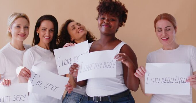 Group of smiling women in white t-shirts holding empowering handwritten signs and dancing together. Happy multiracial young females expressing self love, body positivity, freedom and individuality.