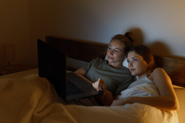 Middle age woman and younger woman relaxing in bed together, watching laptop, promoting active...