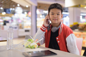 Teen Thai boy wearing headphones eating salad in cafeteria while taking on phone