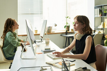 Two young adult women, one Caucasian and one Black, working at computers in modern office,...