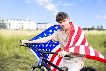 Cheerful teenage boy on a bicycle with an American flag on his shoulders, festive