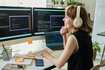Caucasian young adult woman wearing headphones working on neural network development at computer workstation, smiling while coding on multiple monitors with programming interface visible