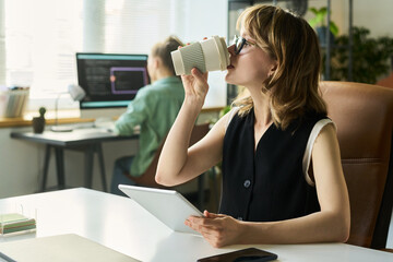 Caucasian young adult woman holding digital tablet and drinking coffee while sitting at desk in...