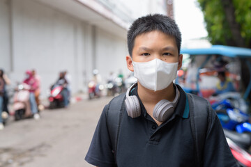 Thai boy with facial mask with rickshaw taxis in background wearing headphones