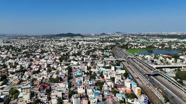 Aerial drone footage of a cityscape in Chennai, India, on a clear, sunny day. Over a multi-lane flyover bustling with vehicle traffic, including cars, trucks, and buses. Parallel to the flyover train