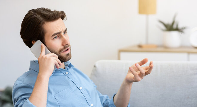 Discontented Guy Frowning Talking On Cellphone Sitting With Laptop, Having Computer Or Internet Connection Problem At Home. Displeased Customer Calling Hotline. Remote Work Issue. Selective Focus