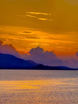 A dramatic sunset paints the sky in fiery oranges and yellows, with soft purple and blue clouds hovering above distant, silhouetted mountains. The vast expanse of the ocean reflects the vibrant hues
