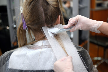 A hairdresser dyes the hair roots light with a brush for a young woman in a barbershop. The hair dye is applied by the hairdresser with a brush.