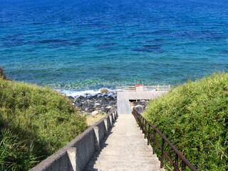 Obraz premium A charming stone stairs leads down to a beautiful Sicilian beach, framed by lush Mediterranean greenery and bathed in warm sunlight. The steps invite visitors to explore the tranquil coastline