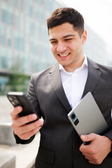 Businessman smiling while checking smartphone and holding tablet outdoors in a modern urban setting...