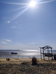 Rustic low‑tide beach scene with a solitary fishing boat and skeletal wooden shelter under a bright midday sun