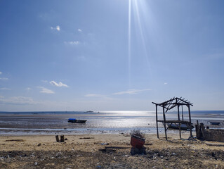 Rustic low‑tide beach with a small fishing boat and skeletal wooden shelter under bright midday sun