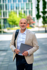 Middle aged man walking on the city street and holding a notebook compute in his hand