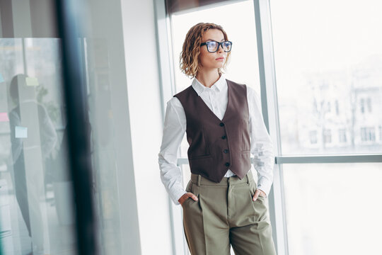 Successful businesswoman in formal attire standing in a bright office with large windows exuding professionalism and style