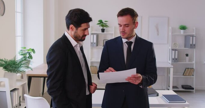 Two business people in suits standing in office, closely reviewing and discussing some data on sheets. Male colleagues or partners working with documents together, collaborating on plans and strategy.