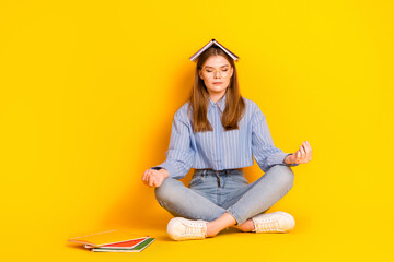 Young woman meditating with book on head in casual clothes seated on yellow backdrop showcasing...