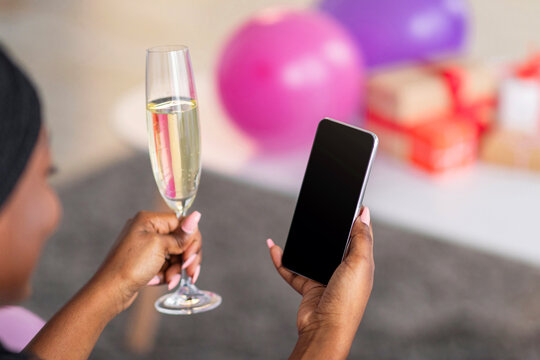 Modern smartphone with blank screen in black lady hand, birthday, valentine, new year online celebration during covid-19 pandemic. African american woman with glass of champagne having video chat - Powered by Adobe