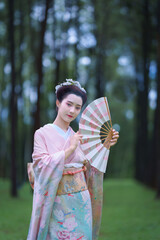 Traditional Japanese  pretty woman in Furisode Kimono holding fan and posting in green natural Pine Forest. Cultural fashion and nature harmony.