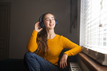 Young woman listening music in wireless headphones smiling sitting alone at home look at window...