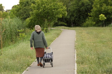 An elderly woman walks along a green avenue with a product cart. The difficult fate of people of declining age. Care for the elderly. Grandma's loneliness