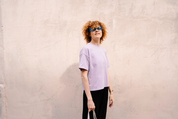 Curly-haired woman in pastel outfit standing against wall