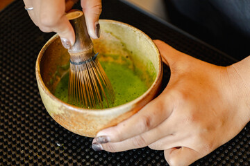 Whisking green matcha tea in rustic ceramic bowl with bamboo whisk, hands gently holding bowl on...