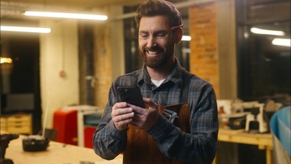 Happy Caucasian man carpenter using mobile phone in workshop wood master craftsman swiping screen...