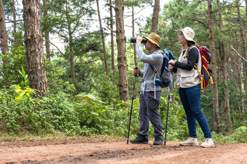 Asian male bird watcher using binocular and female trekker holding poles trekking in national forest